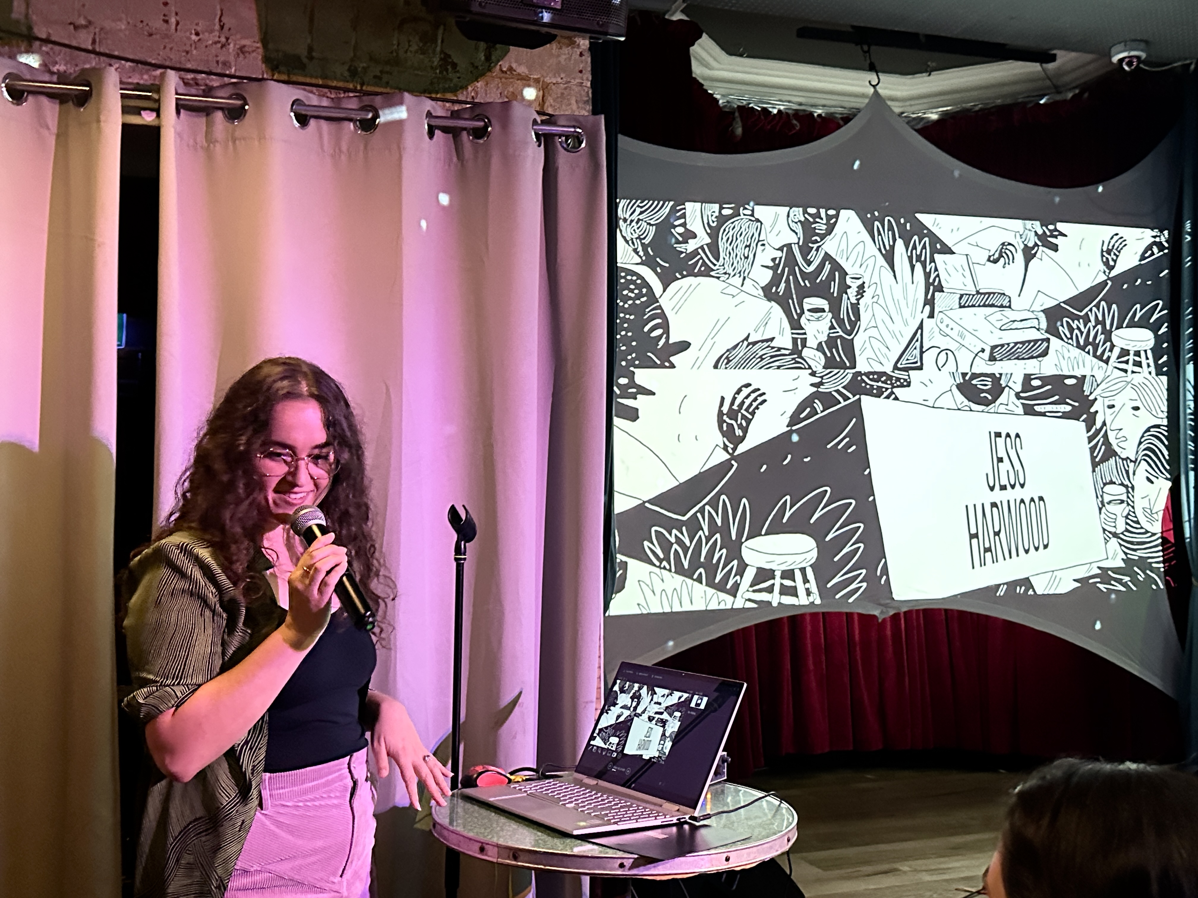 A woman with long brown hair holds a microphone in front of a projected image saying &quot;Jessica Harwood&quot;